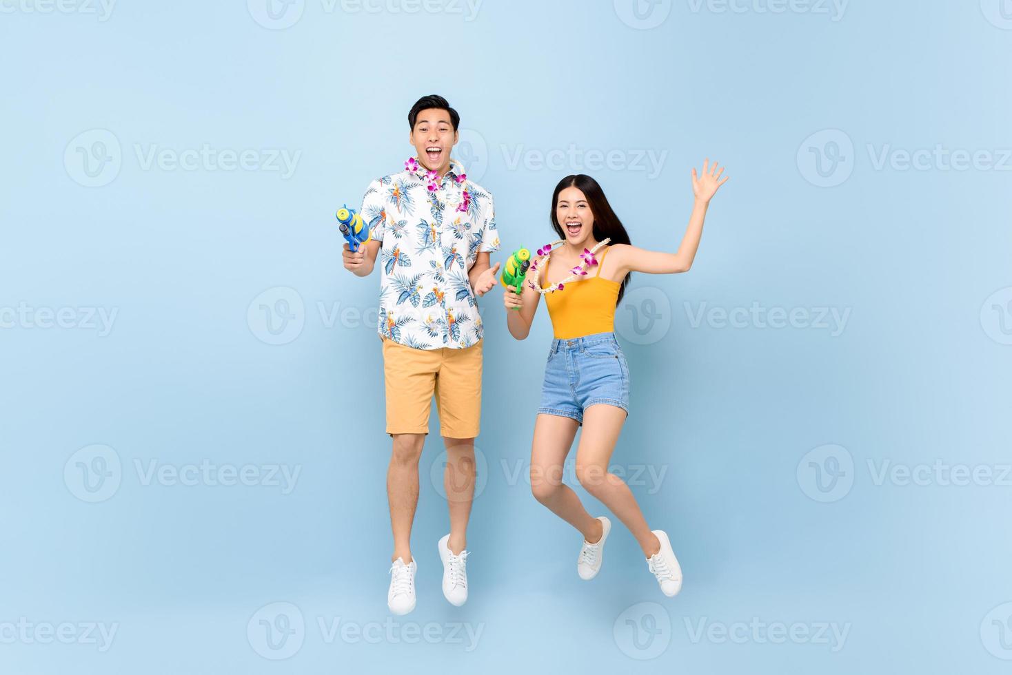 Young Asian couple in summer outfits with water guns jumping in studio blue background for Songkran festival in Thailand and southeast Asia photo