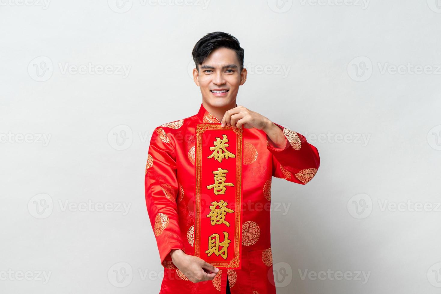 Handsome smiling Asian man showing red label with text means may you attain greater wealth in isolated studio light gray background for Chinese new year concepts photo
