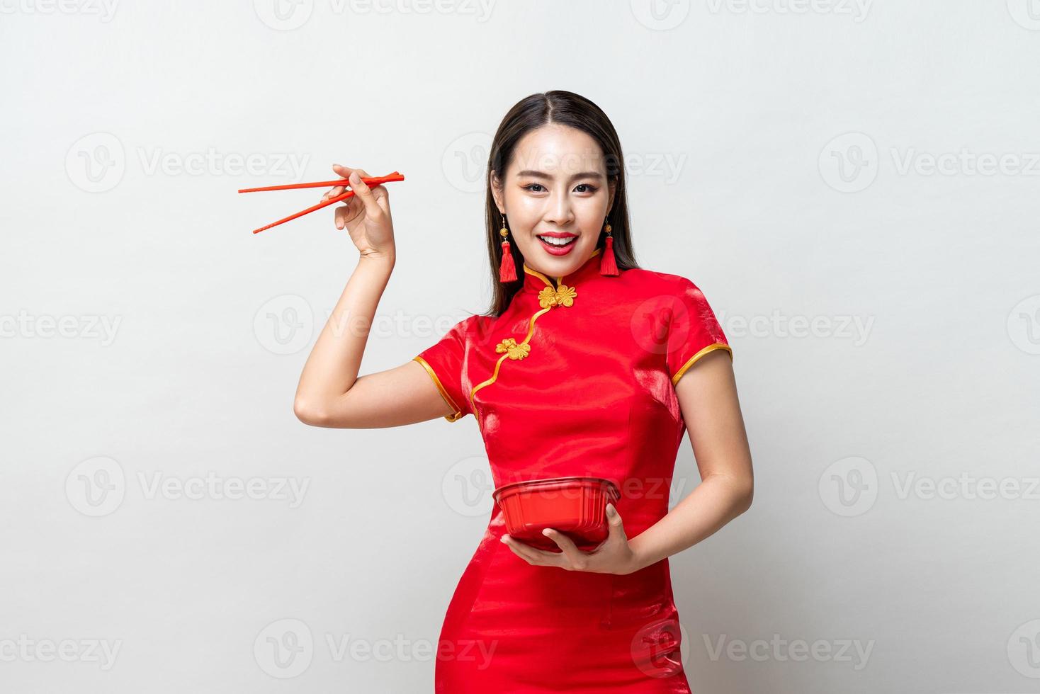 Pretty Asian woman in traditional red Chinese qipao dress holding plastic food box and chopsticks in studio isolated gray background photo