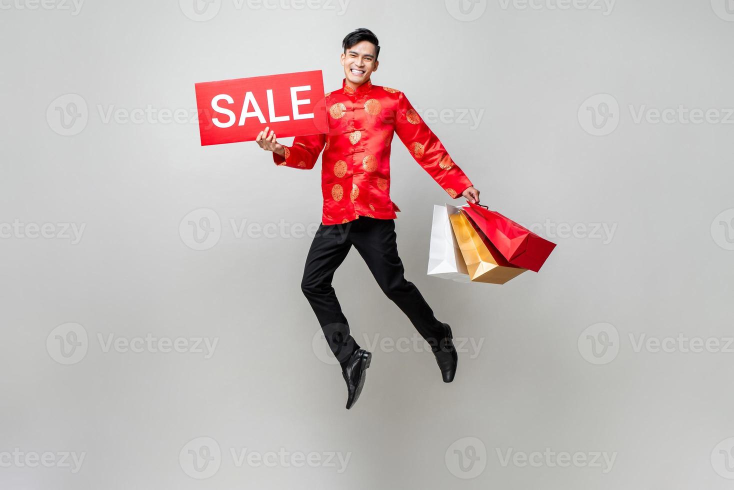 Surprised Asian man in traditional costume holding bags and red sale sign jumping in isolated light gray background for Chinese new year shopping concept photo