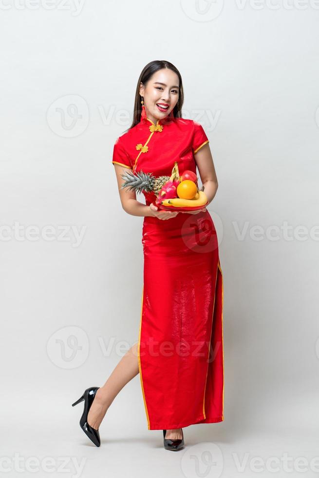 Young Asian woman in traditinal red cheongsam dress holding lucky fruits for Chinese new year in light gray isolated studio background photo