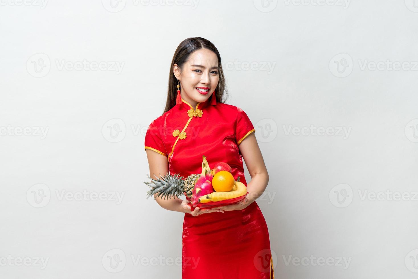 Young Asian woman in traditinal red cheongsam dress holding lucky fruits for Chinese new year in light gray isolated studio background photo