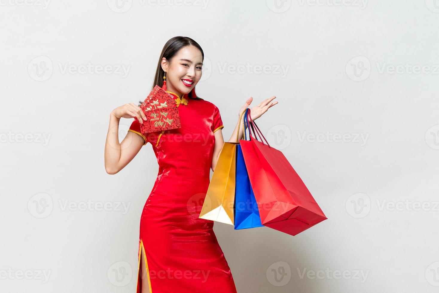 Asian woman in traditional dress holding bags and red envelopes in isolated light gray studio background for Chinese new year shopping concept, text means great luck great profit photo