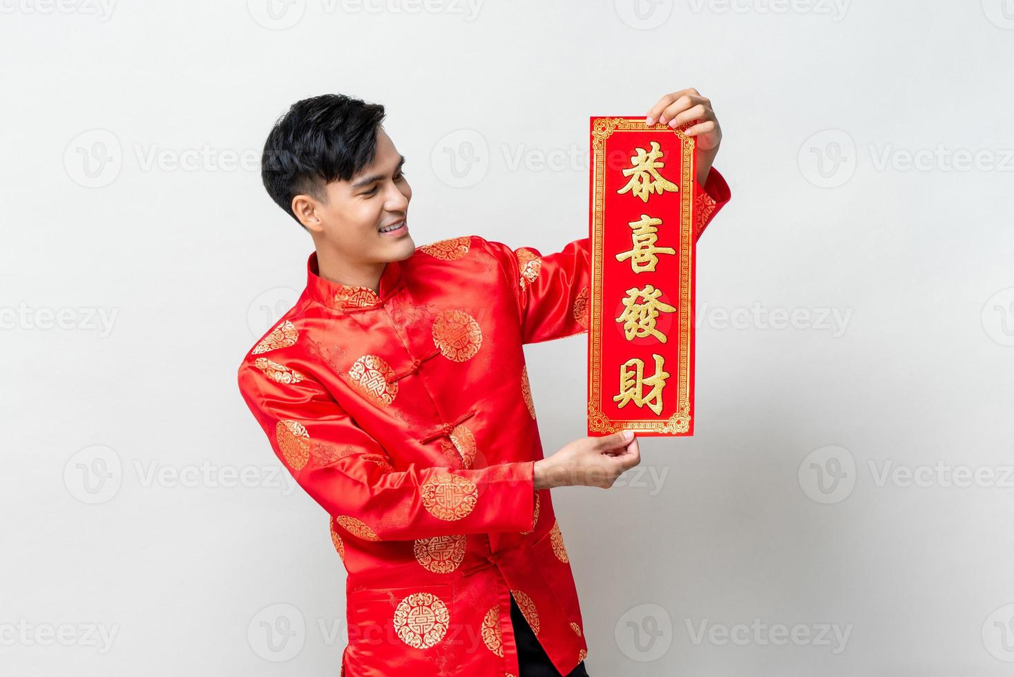 Handsome smiling Asian man showing red label in isolated studio light gray background for Chinese new year concepts, text means may you attain greater wealth photo