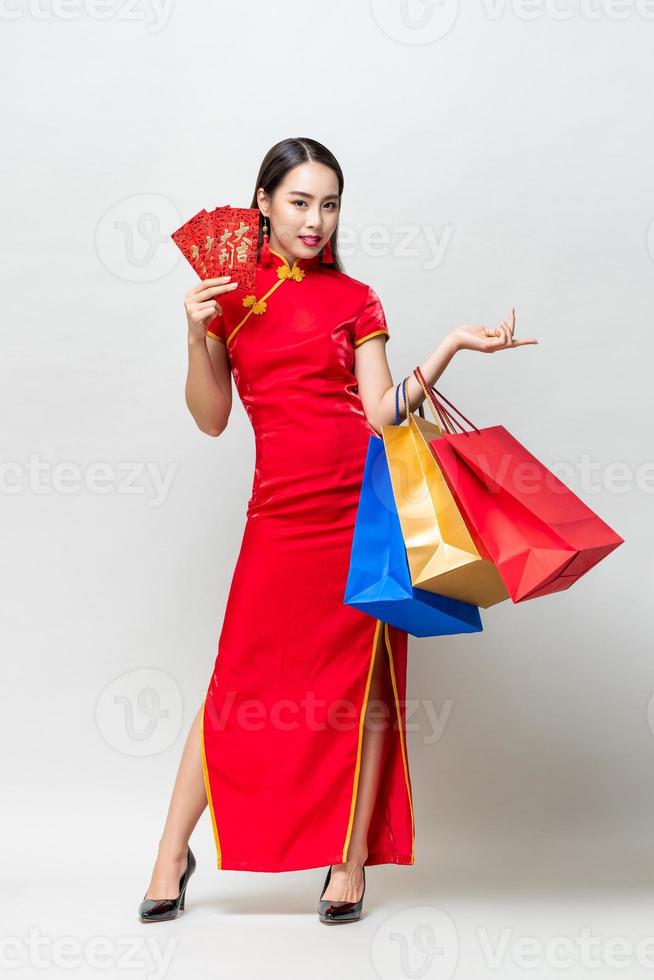 mujer asiática con vestido tradicional sosteniendo bolsas y sobres rojos en un estudio gris claro aislado para el concepto de compras de año nuevo chino, el texto significa gran suerte gran beneficio foto