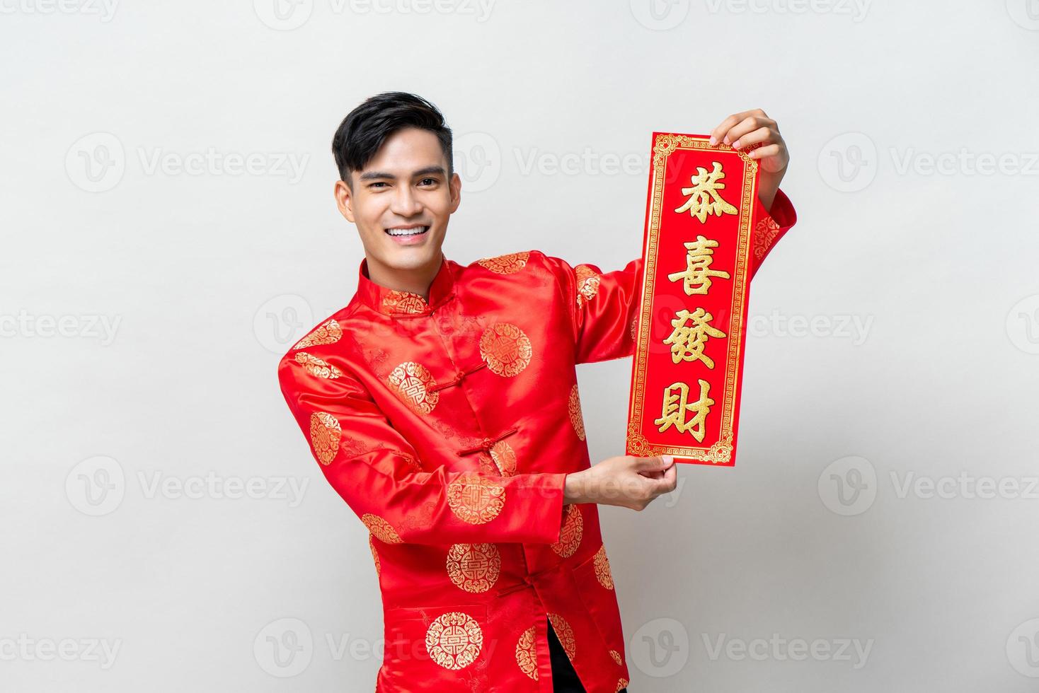 Handsome smiling Asian man showing red label with text means may you attain greater wealth in isolated studio light gray background for Chinese new year concepts photo