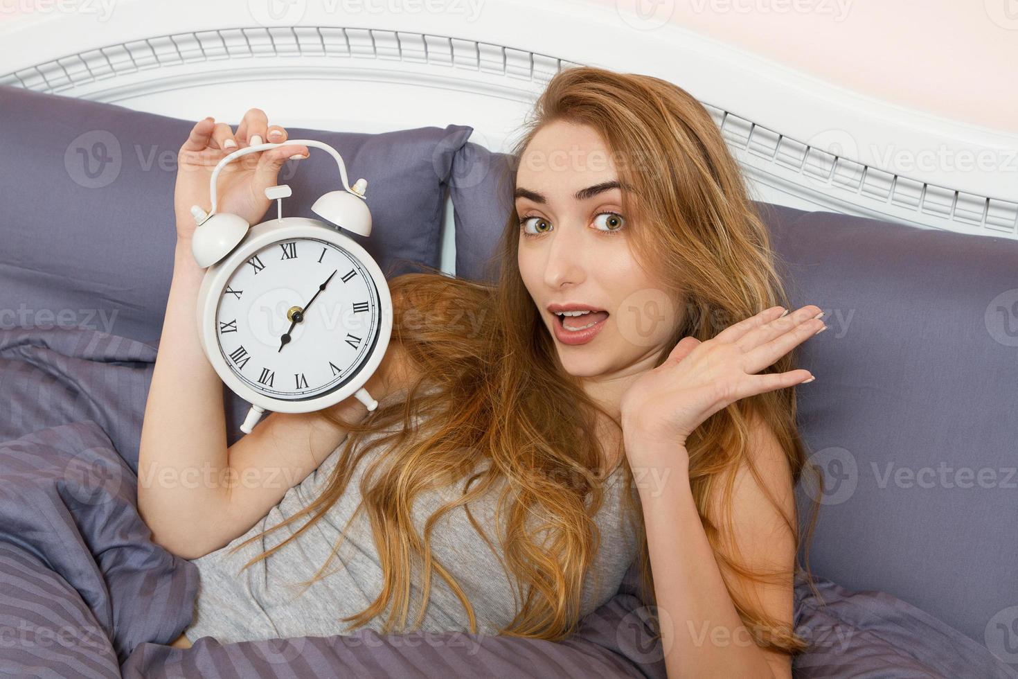 hermosa chica sostiene un reloj y se encuentra en el dormitorio de la cama. hermosa mujer emocional acostada en la cama en su día libre. foto