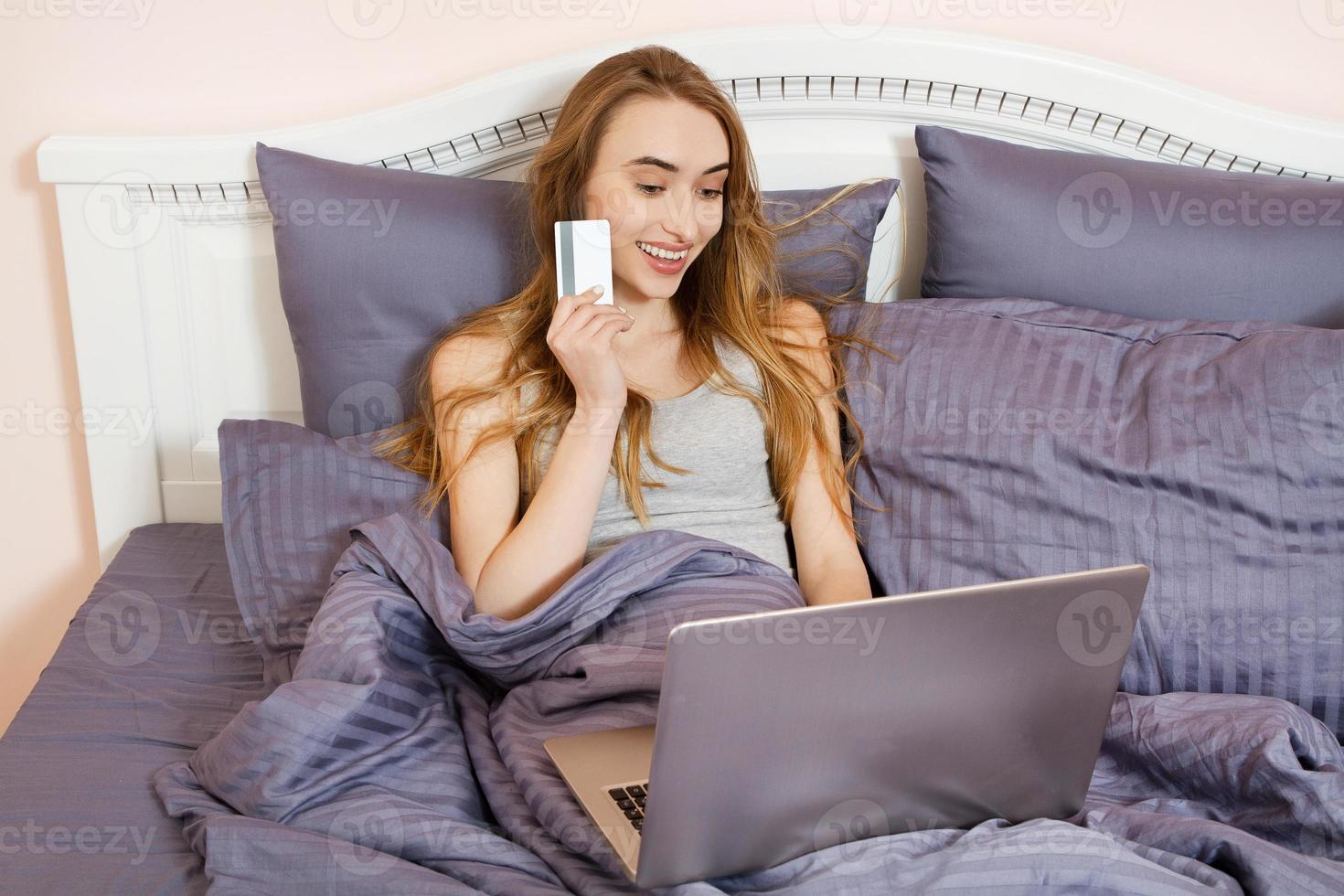 mujer que usa una computadora portátil para comprar en la tienda en línea en la cama en casa, pago con tarjeta de crédito con transacciones bancarias en línea. niña feliz señaló en tarjeta de crédito. foto
