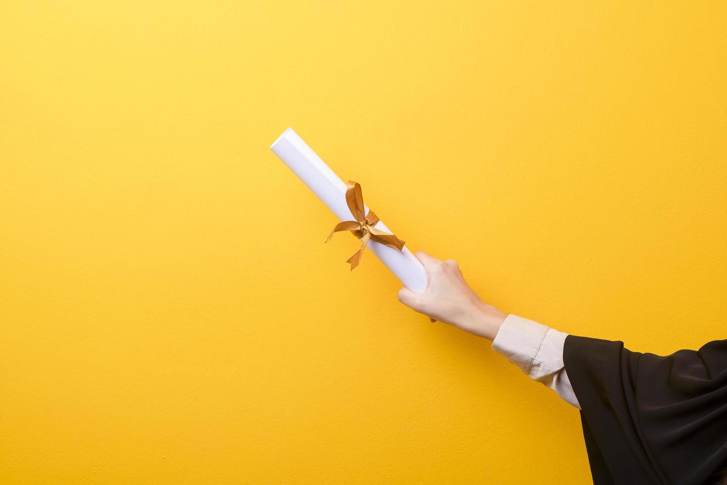 Close up of woman hand in graduation gown is holding graduation cap and certificate on yellow background photo