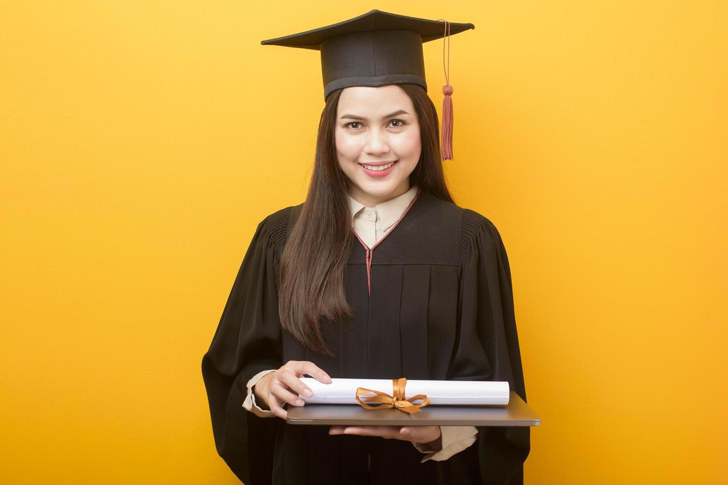portrait of beautiful woman in graduation gown is holding laptop computer on yellow background photo