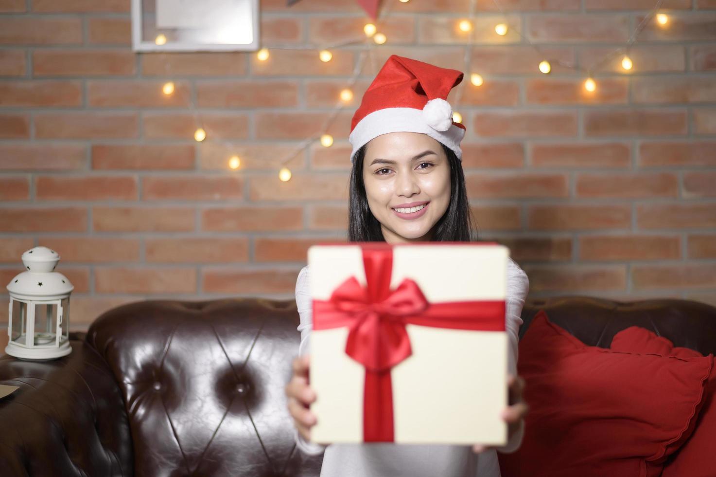 Young smiling woman wearing red Santa Claus hat showing a gift box on Christmas day, holiday concept. photo