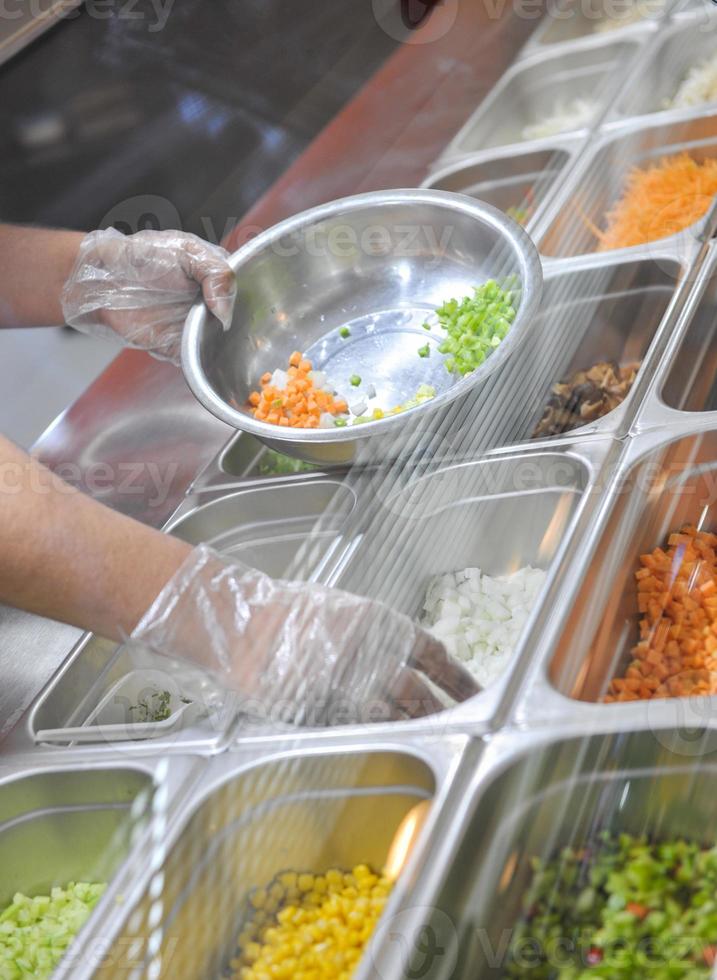 el cocinero pone trozos de verduras para ensalada en un bol. bandeja con ensalada variada en la ventana de un restaurante de comida rápida foto