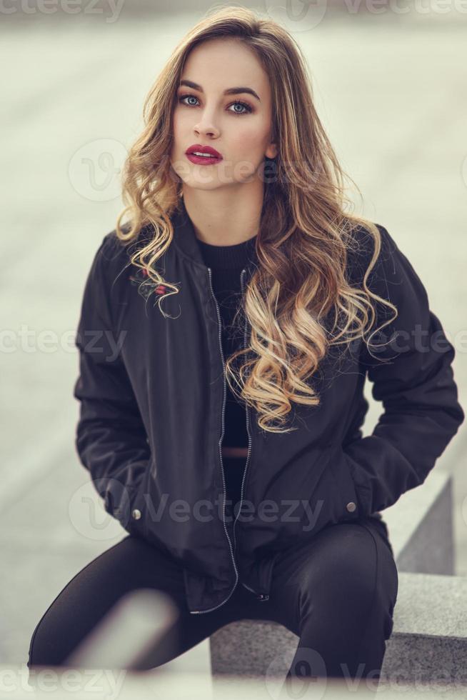 Beautiful young girl wearing black jacket sitting in the street. photo