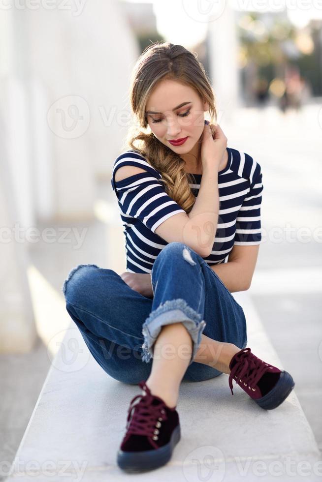 Blonde woman, model of fashion, sitting in urban background. photo