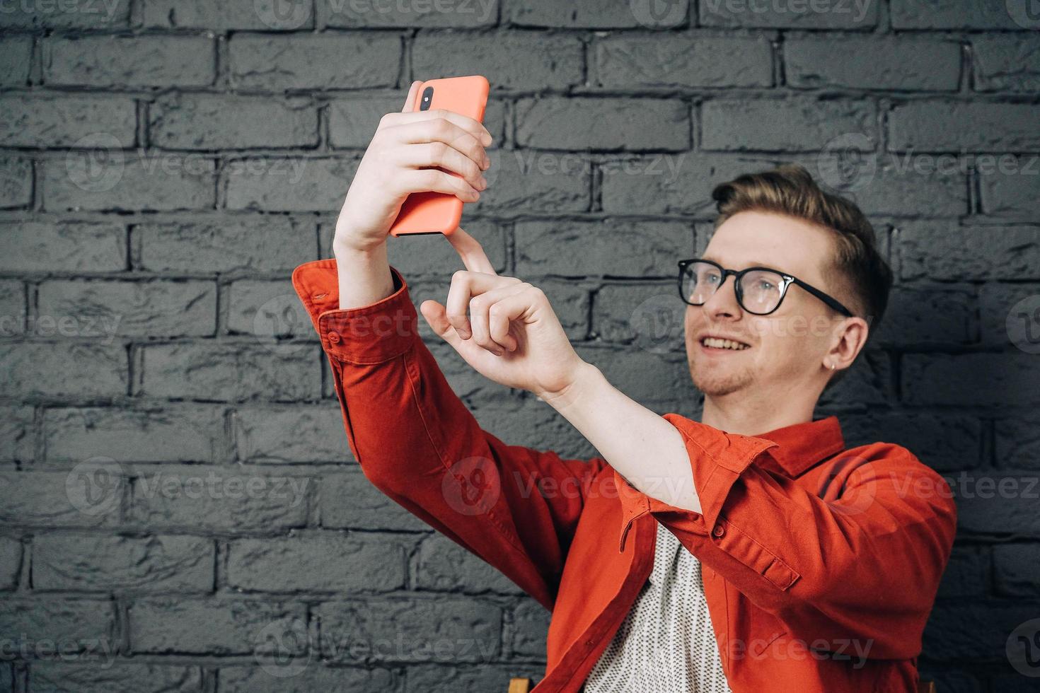 Young joyful man in red shirt and glasses taking selfie photo on cellphone on a background of black brick wall. Copy, empty space for text