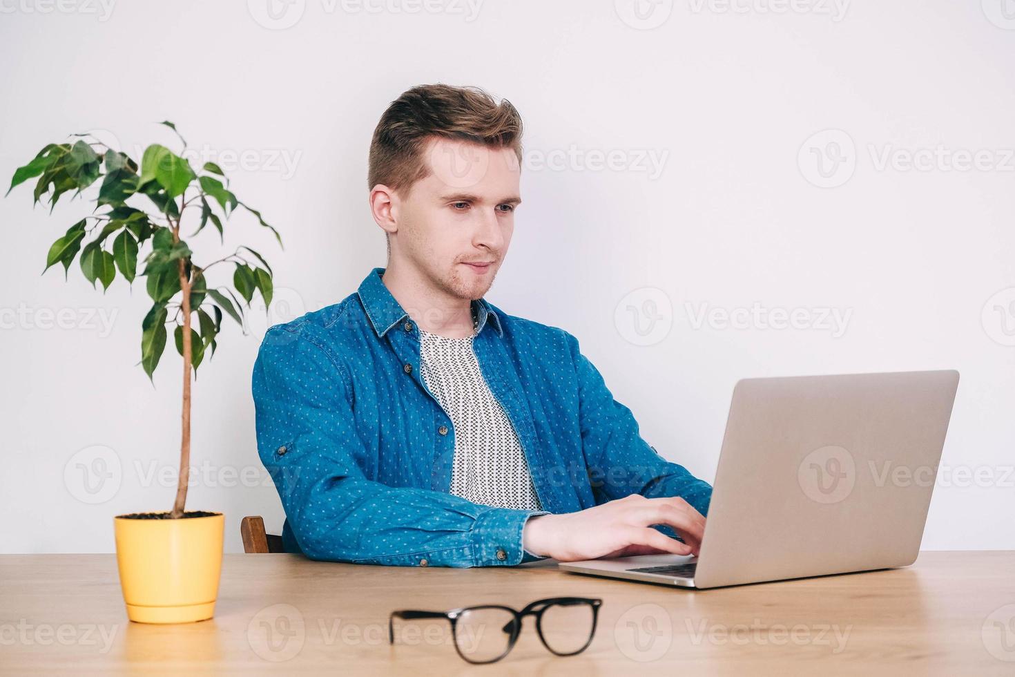 Young man in shirt and glasses working on laptop computer sitting at the table on a white background. Freelance work concept. Copy, empty space for text photo