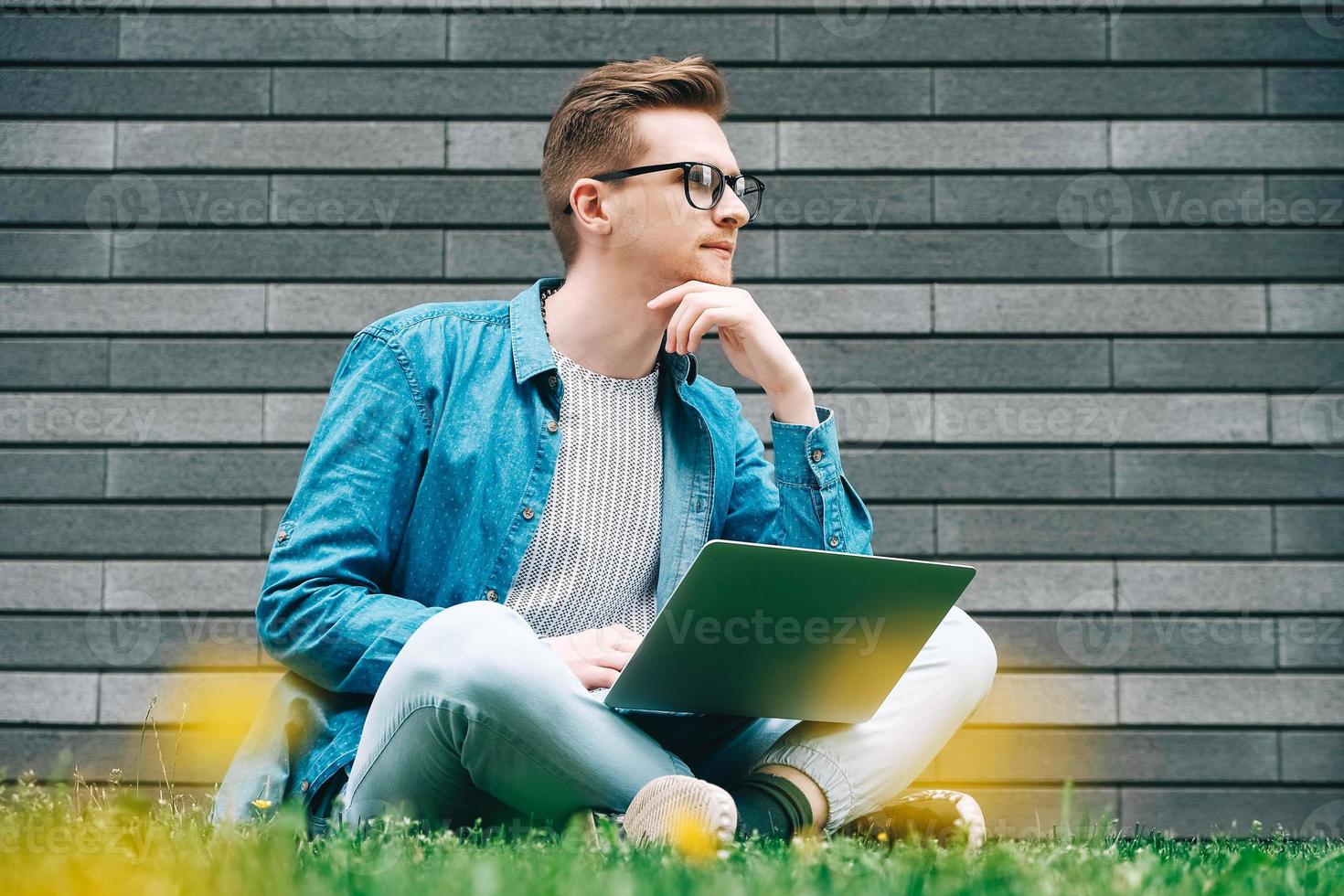 hombre con anteojos hombre de negocios hipster sentado en la hierba verde y usando una computadora portátil en un fondo de pared gris foto