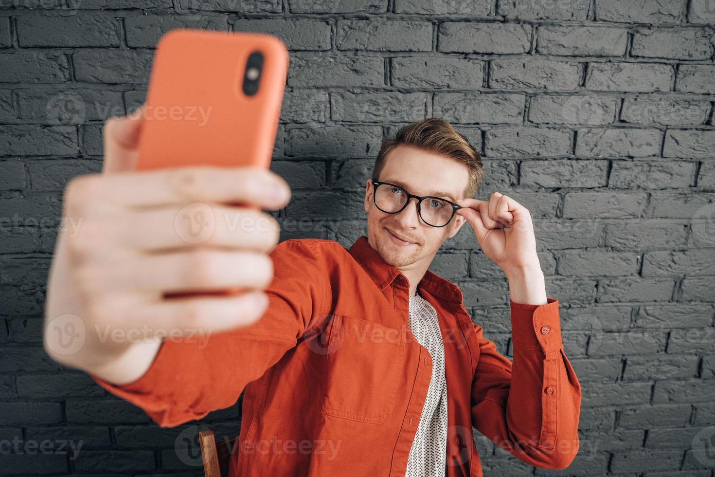 Young joyful man in red shirt and glasses taking selfie photo on cellphone on a background of black brick wall. Copy, empty space for text