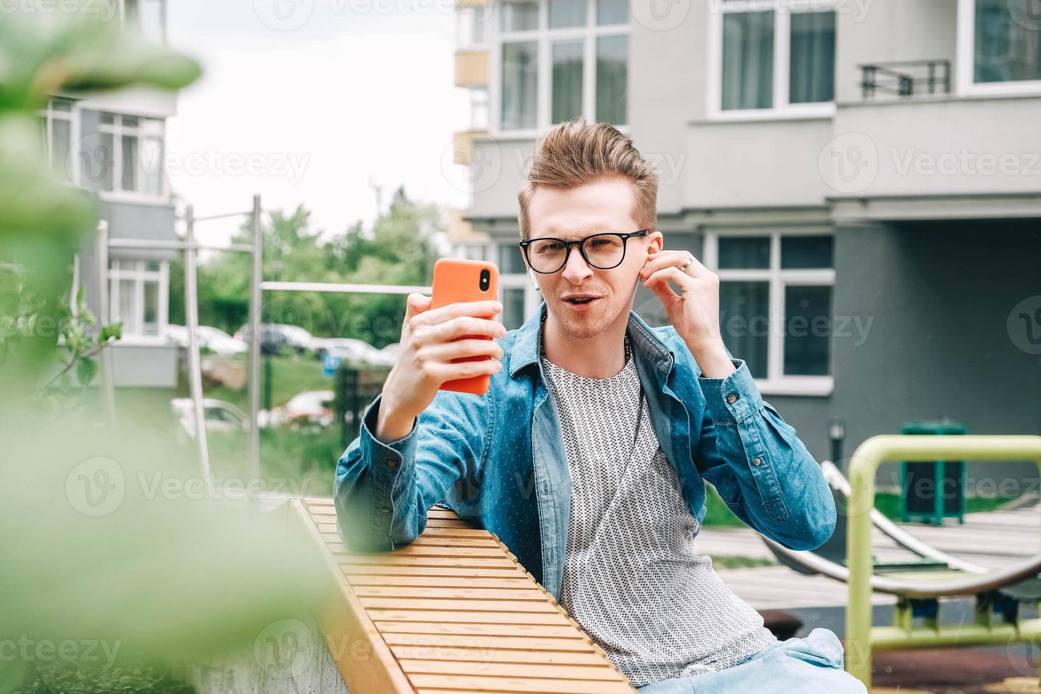 Portrait man in shirt and glasses using communicates on a smartphone sitting on a bench on a background of houses photo