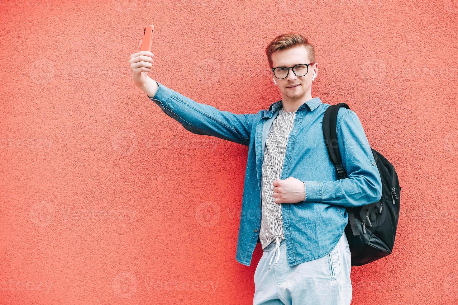 Man in shirt, jeans and glasses carrying a backpack and looking at smartphone while standing on a pink background photo