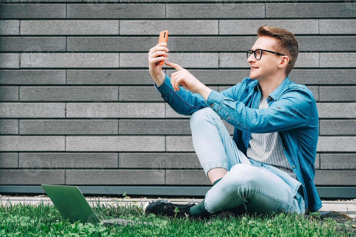 Man with glasses sitting on green grass and looking at smartphone on a gray wall background photo
