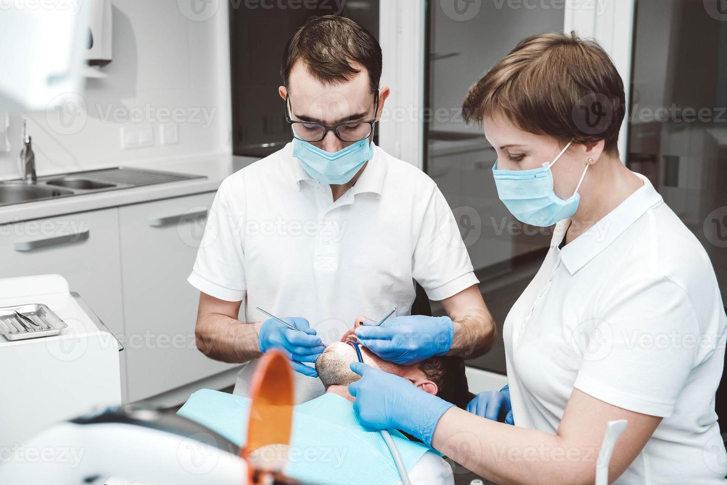 el dentista y su asistente tratan a un paciente masculino en una clínica dental. dentistas con máscaras médicas trabajando con los dientes del cliente foto