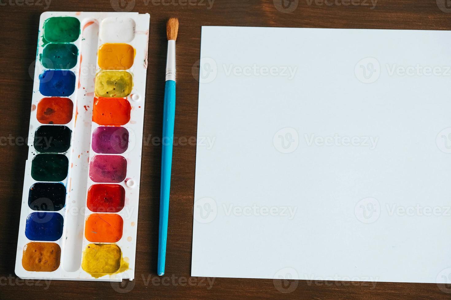 Set of watercolor paints, brush for painting and blank white paper sheet on wooden background photo