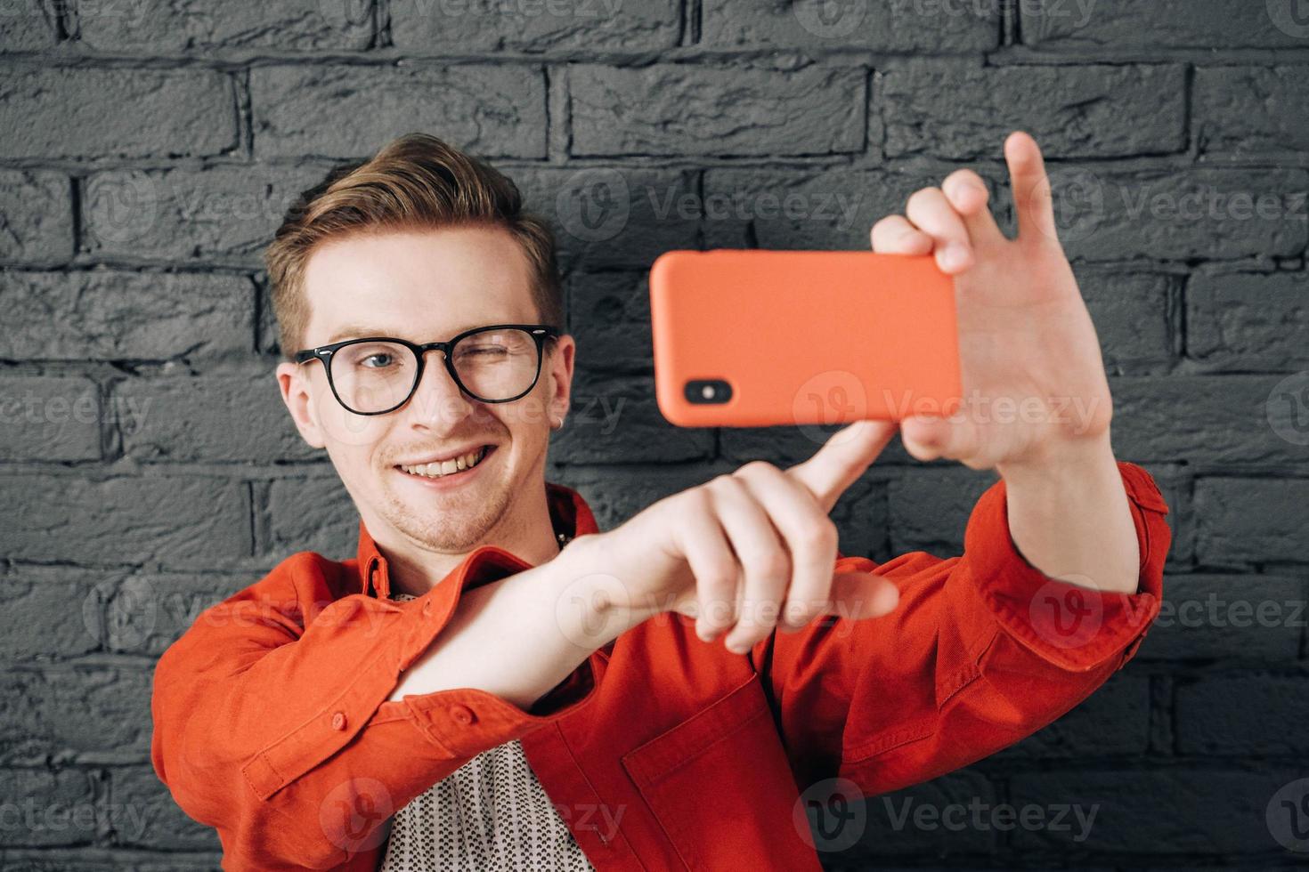 joven alegre con camisa roja y gafas tomando una foto selfie en el celular sobre un fondo de pared de ladrillo negro. copiar, espacio vacío para texto