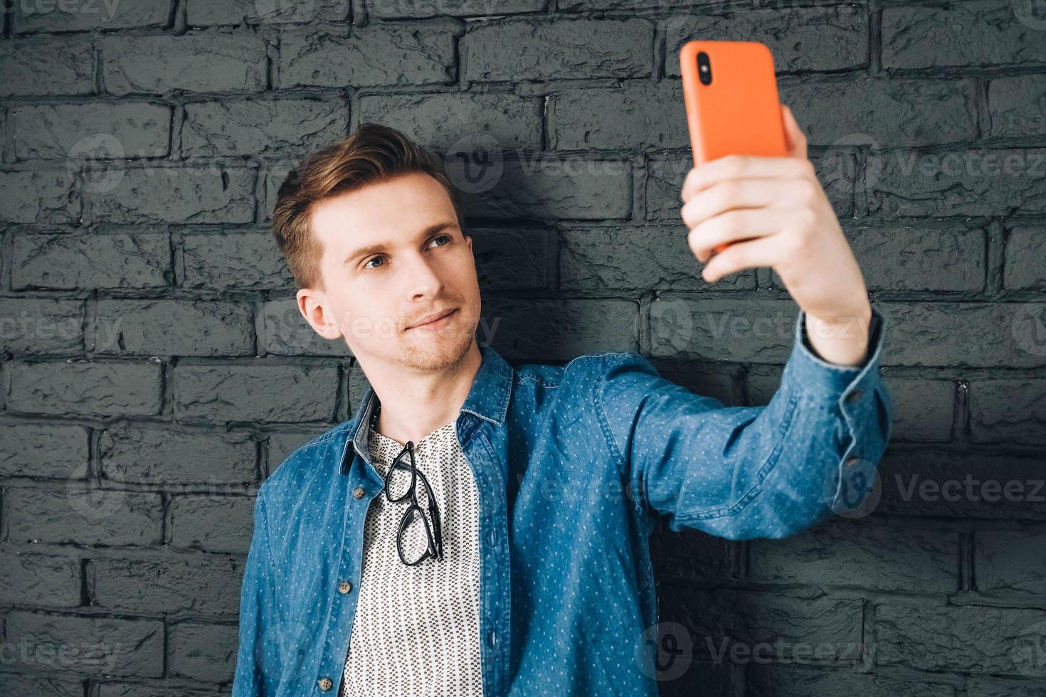 Young joyful man in blue shirt and glasses taking selfie photo on cellphone on a background of black brick wall. Copy, empty space for text