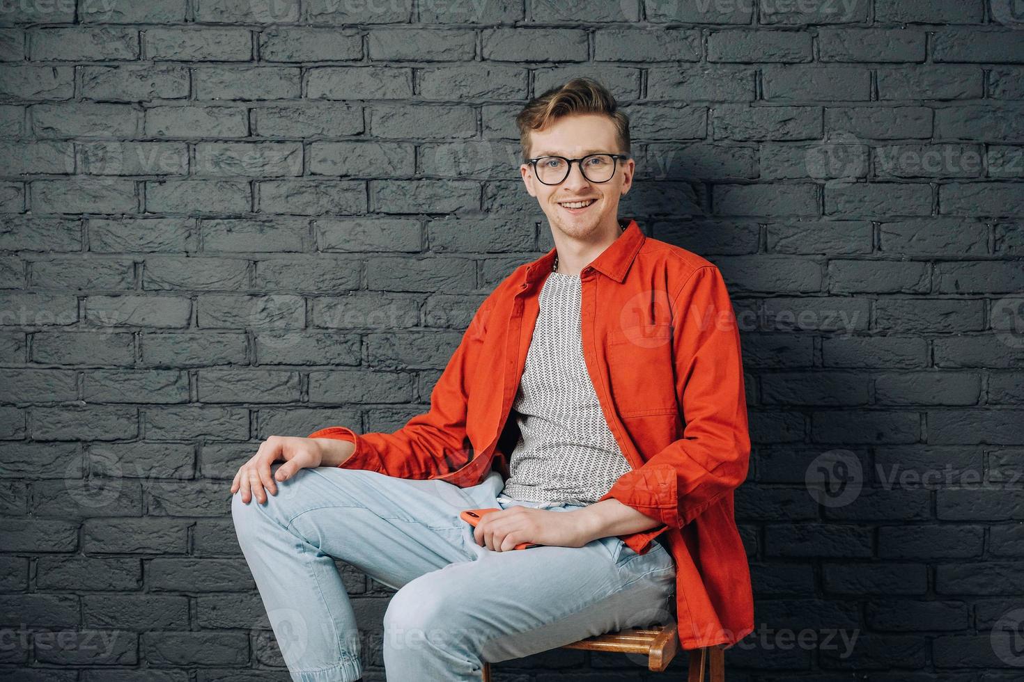 Young joyful man in red shirt and glasses holding smartphone sitting on chair on a background of black brick wall. Copy, empty space for text photo