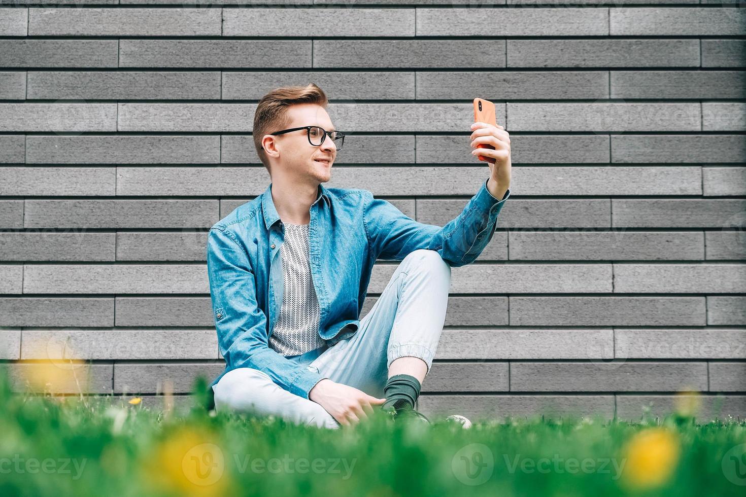 Man with glasses sitting on green grass and looking at smartphone on a gray wall background photo