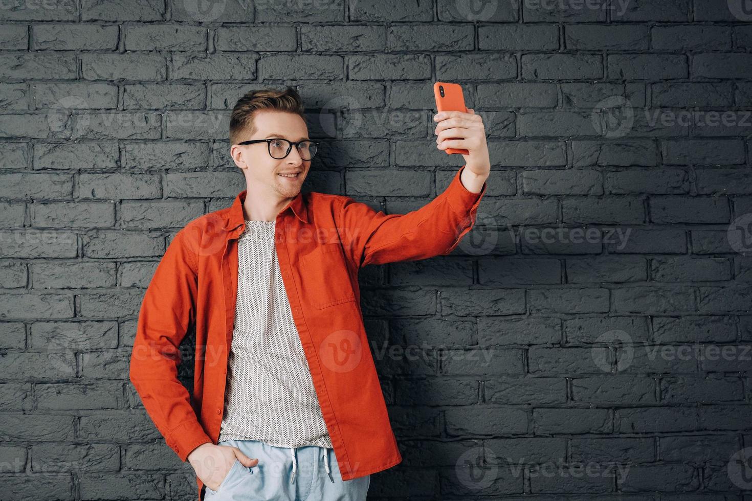 Young joyful man in red shirt and glasses taking selfie photo on cellphone on a background of black brick wall. Copy, empty space for text