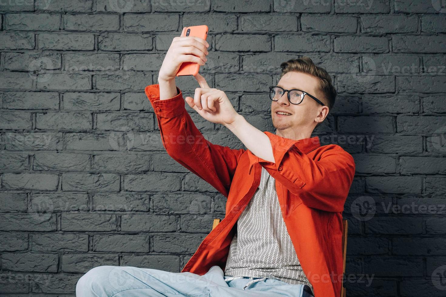 Young joyful man in red shirt and glasses taking selfie photo on cellphone on a background of black brick wall. Copy, empty space for text