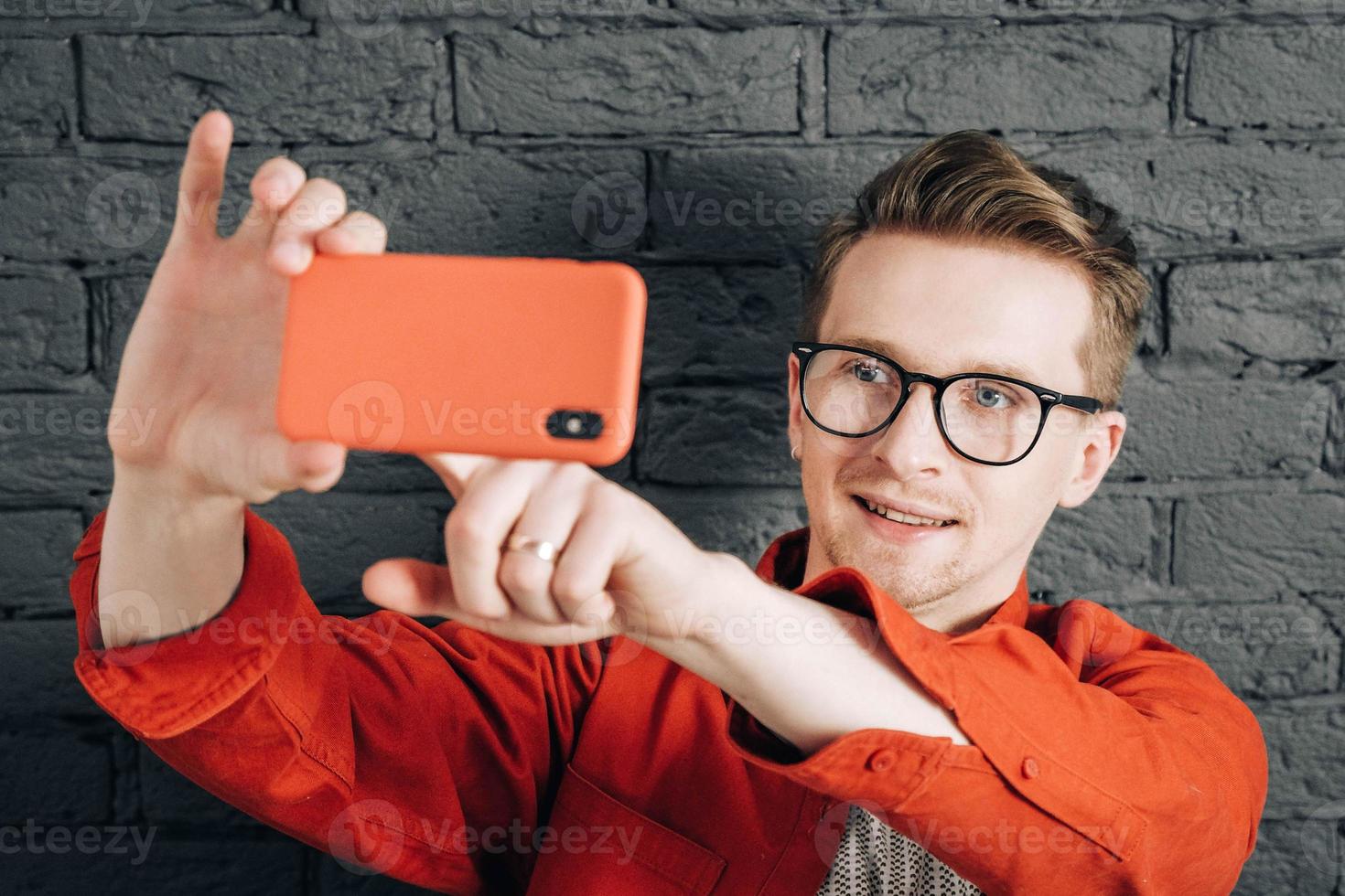 Young joyful man in red shirt and glasses taking selfie photo on cellphone on a background of black brick wall. Copy, empty space for text