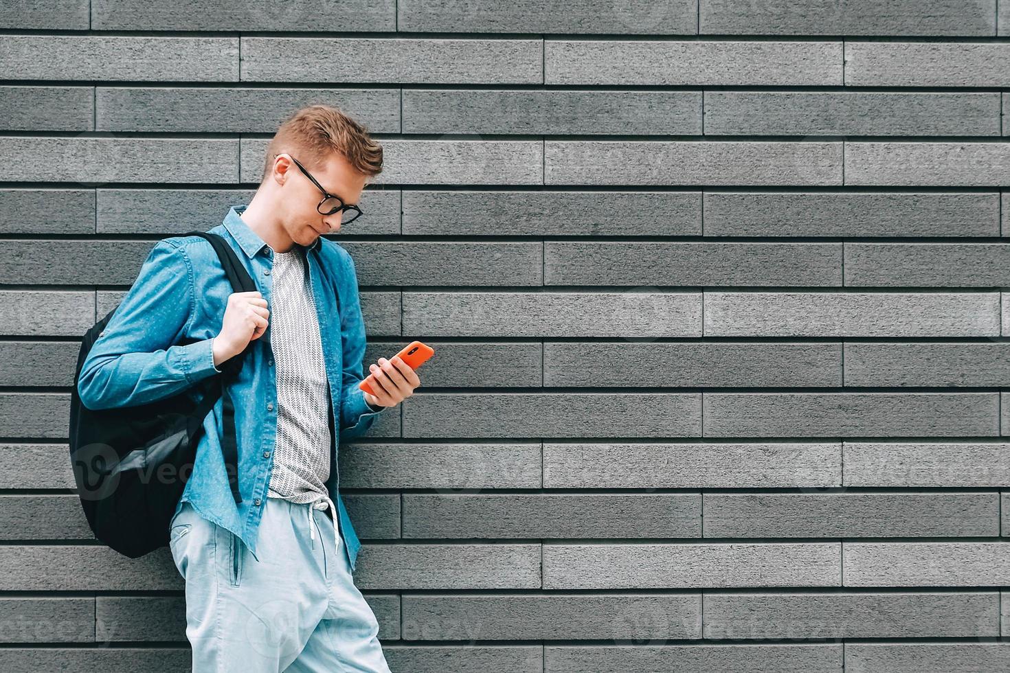 Man in shirt, jeans and glasses carrying a backpack and looking at smartphone while standing on a background of a gray wall photo