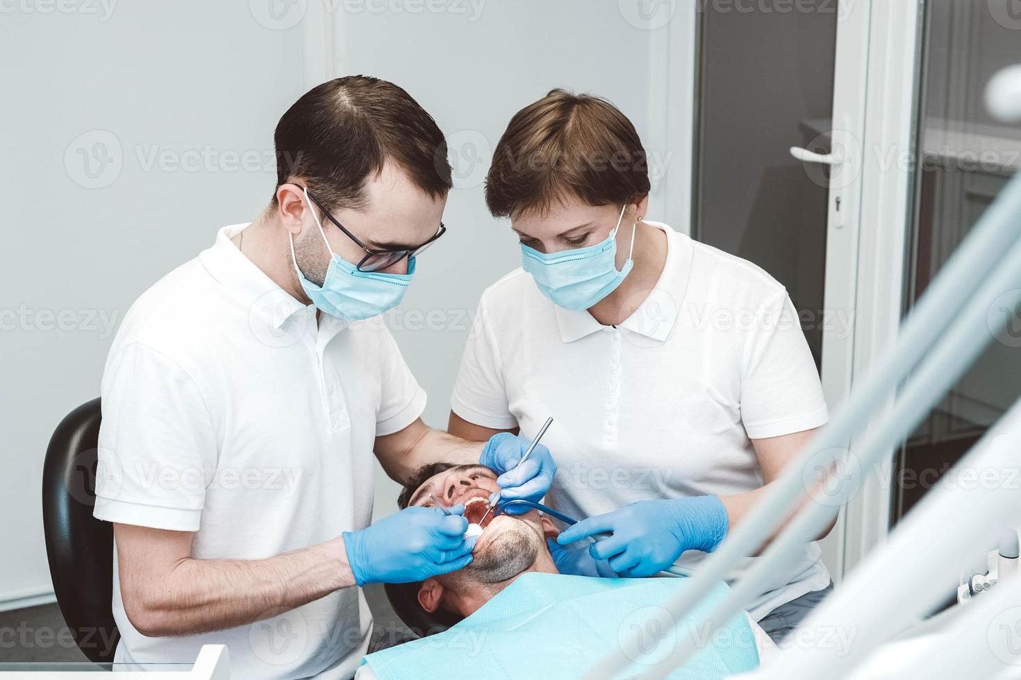 Dentist and his assistant treat a male patient in a dental clinic. Dentists in medical masks working with client's teeth photo