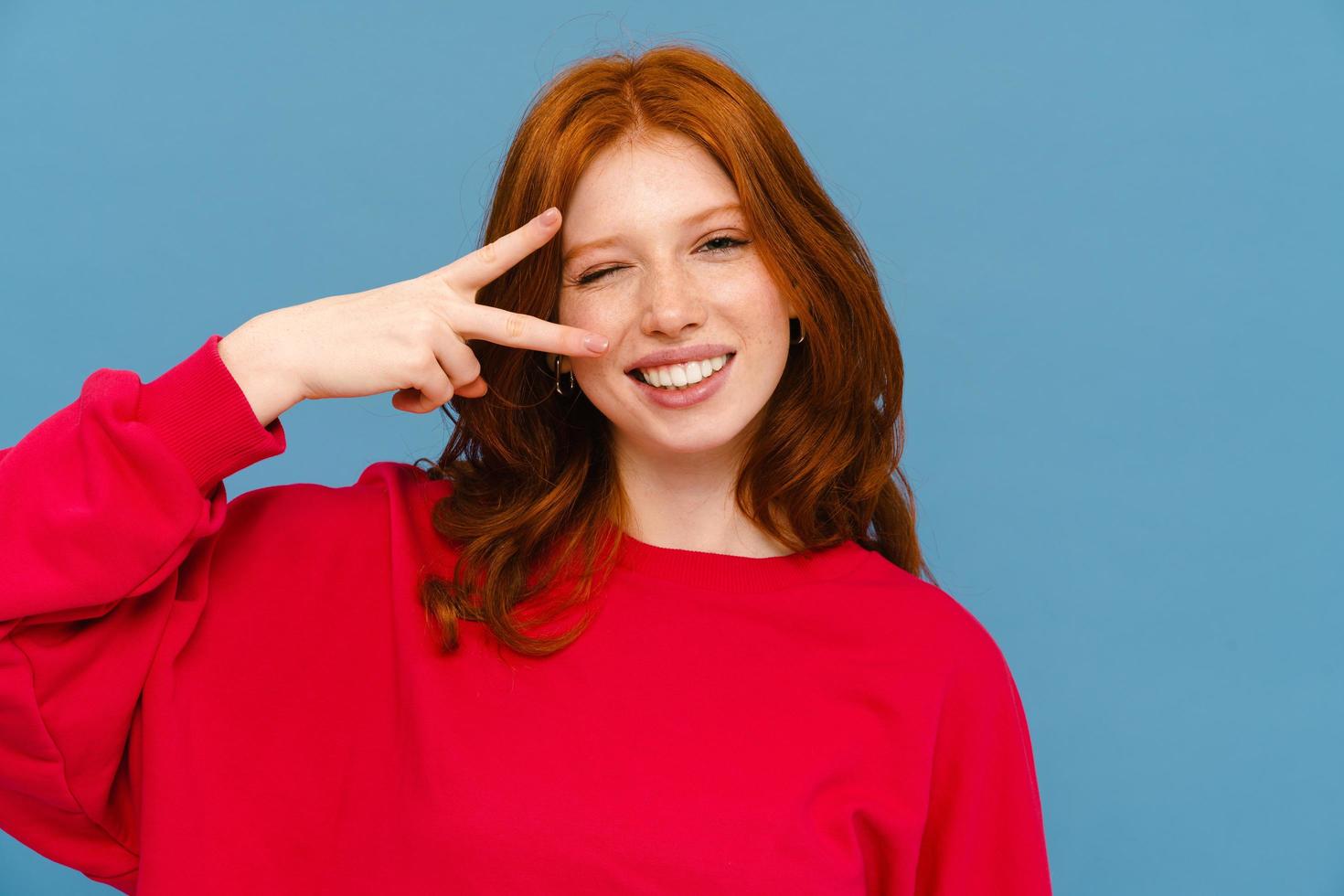 Ginger-haired woman wearing red sweater winking and making peace gesture photo