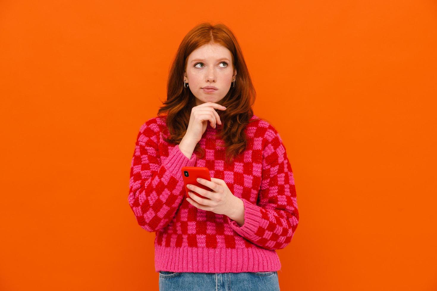 Perplexed ginger-haired woman in plaid sweater using mobile phone photo