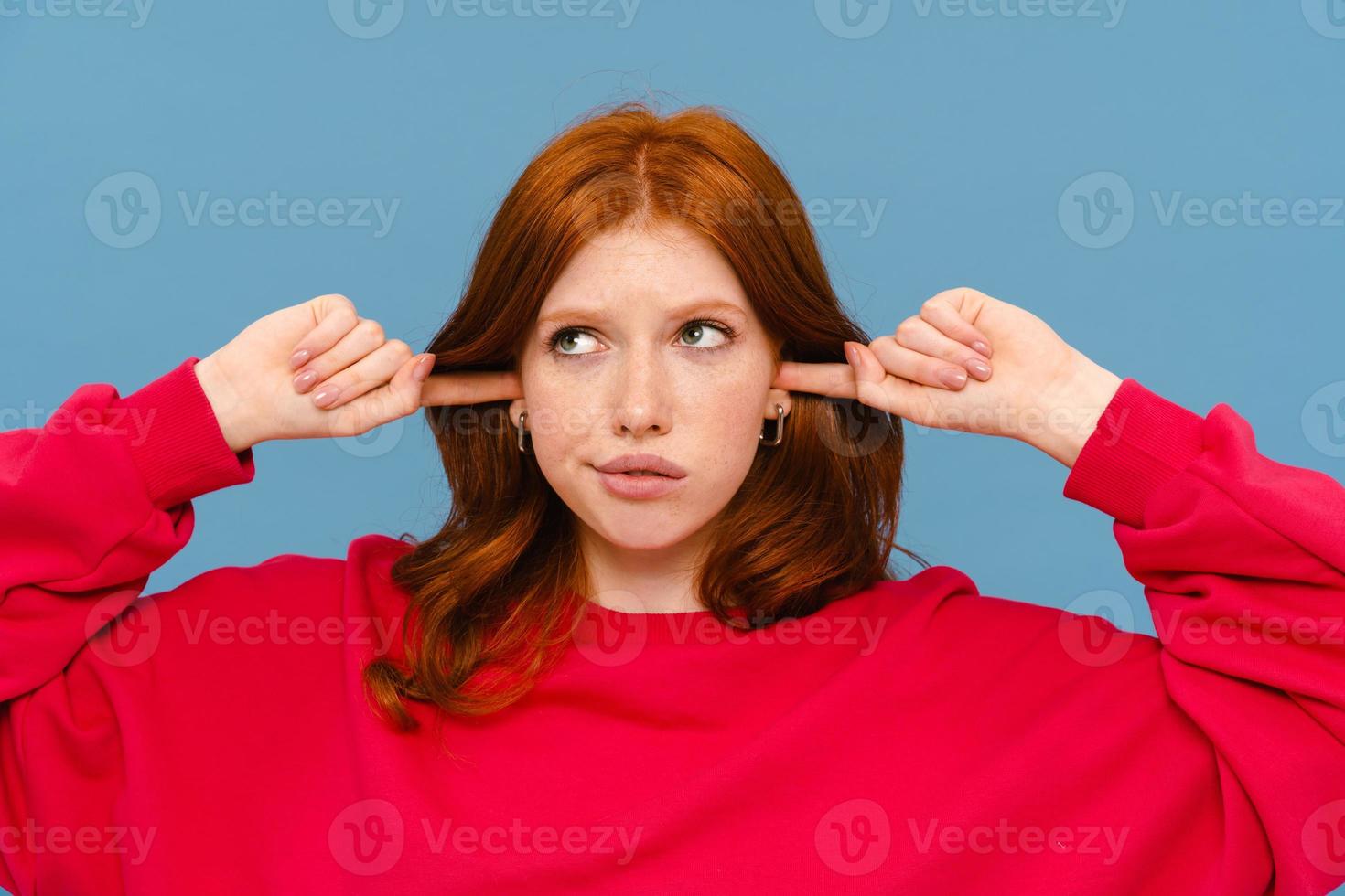 Perplexed ginger-haired woman wearing red sweater plugging her ears photo