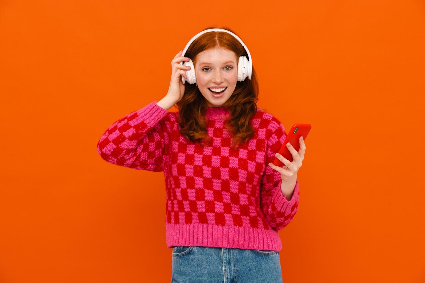 Young smiling ginger-haired woman in plaid sweater using cell phone and headphones photo