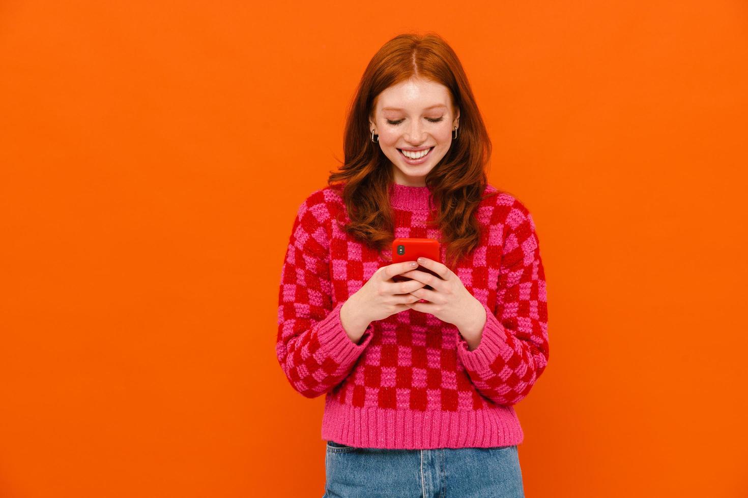 Young ginger-haired woman in plaid sweater smiling and using cellphone photo
