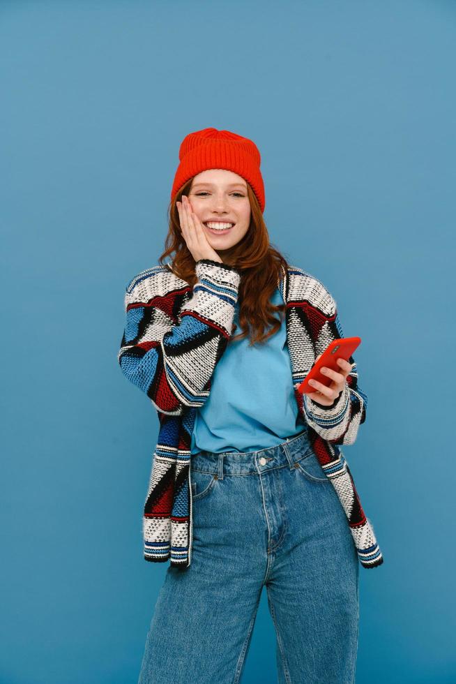 Smiling ginger-haired woman in multicolored sweater using cellphone photo