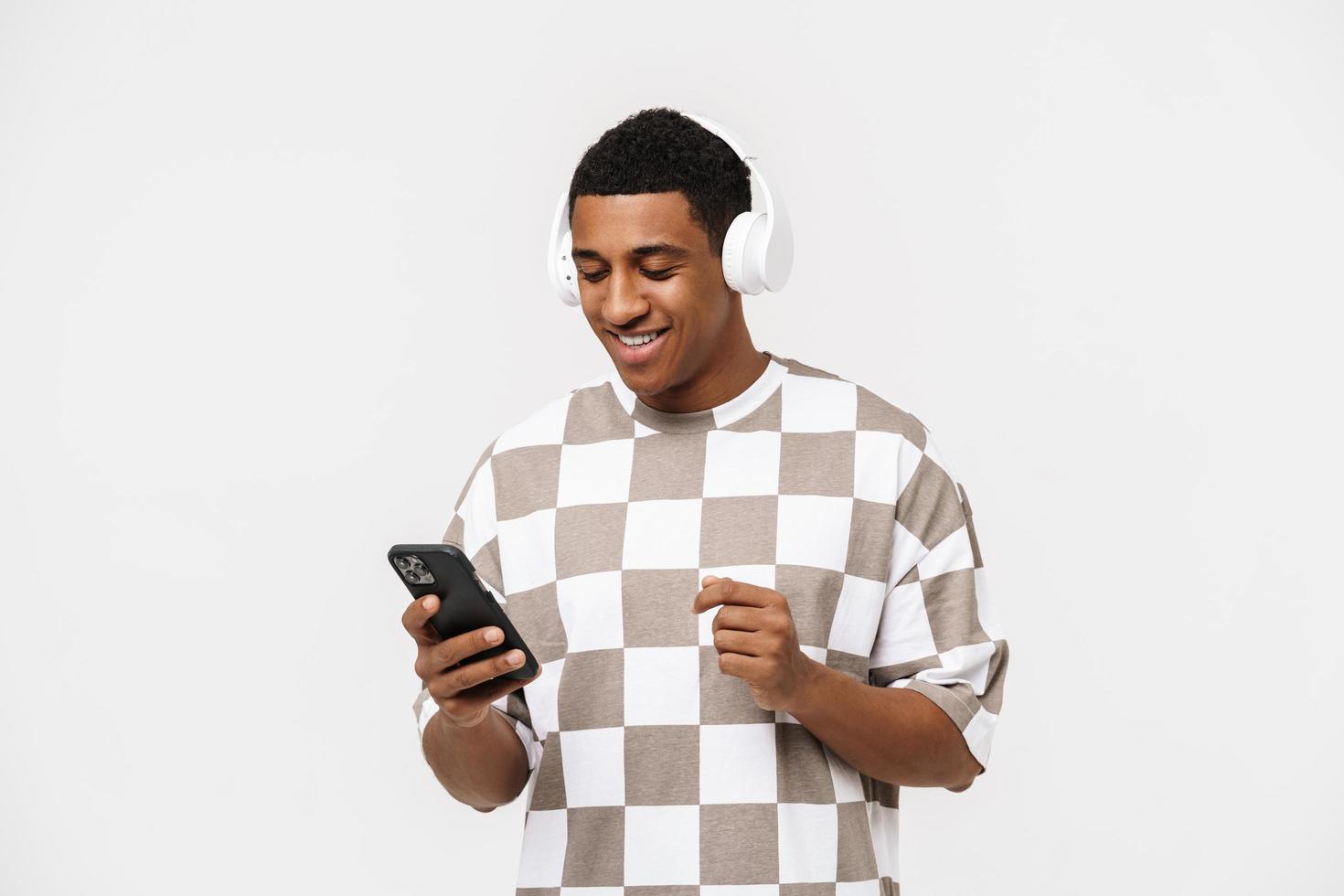 hombre africano alegre mirando el teléfono con auriculares en el estudio blanco foto