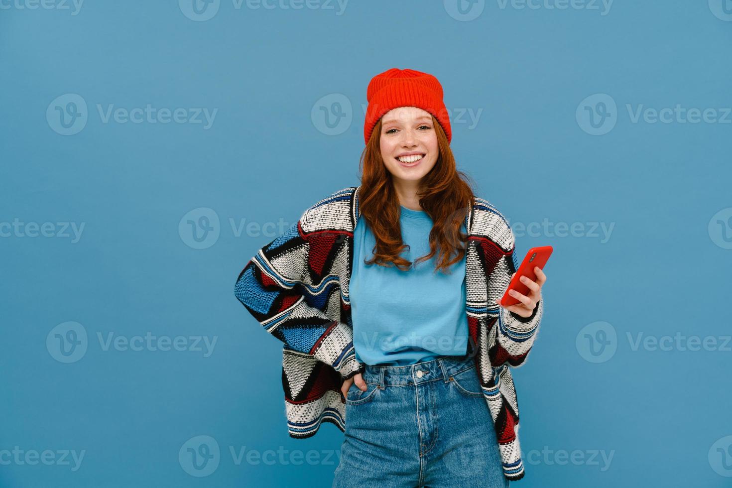 Smiling ginger-haired woman in multicolored sweater using cellphone photo