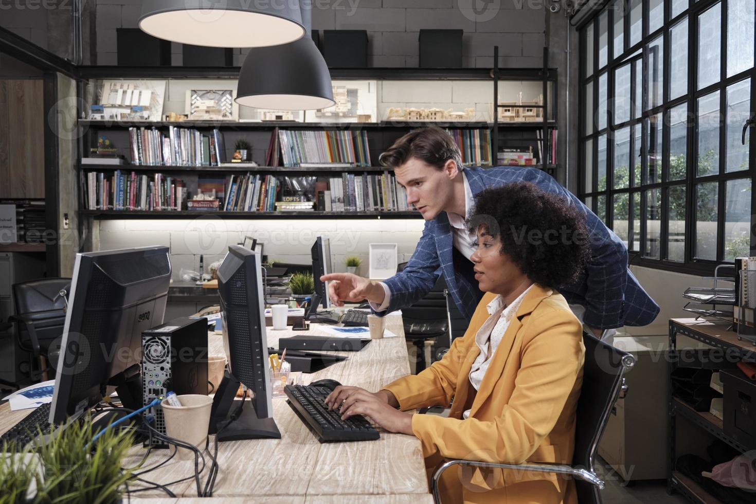 Young African American female worker sits, works with computer, brainstorming, talking, and discussing with Caucasian male colleague and partnership about business jobs in workspace office company. photo