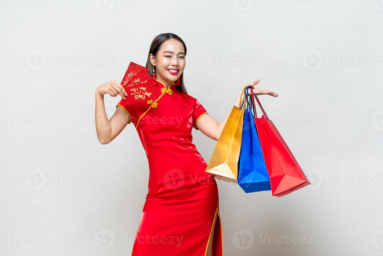 Asian woman in traditional qipao dress holding bags and red envelopes in isolated light gray studio background for Chinese new year shopping concept, text means great luck great profit photo