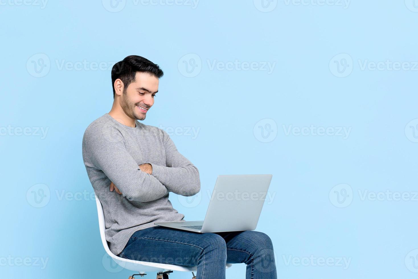 Portrait of smiling happy young Asian man sitting and looking at laptop computer with arms crossed isolated on light blue studio background photo