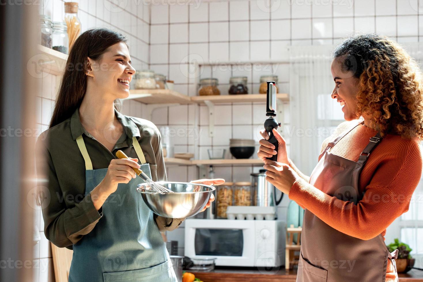 dos amigas felices filmando videos con teléfonos móviles para compartir en línea mientras cocinan en la cocina en casa foto