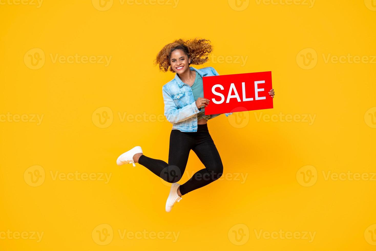 Surprised smiling woman jumping with red sale promotion sign isolated on yellow background photo