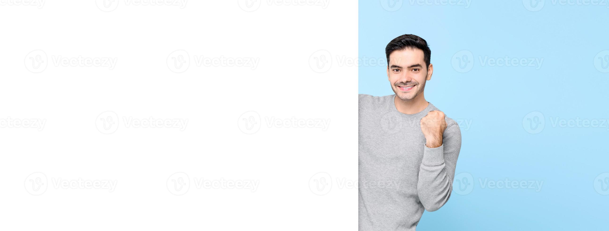 Smiling handsome Caucasian man raising fist behind empty white board on light blue banner background photo